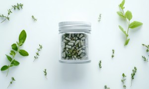 Herbal supplement manufacturing – A transparent jar filled with green herbal supplement capsules, surrounded by fresh green leaves and natural herbs on a white background, emphasizing organic wellness and natural health products