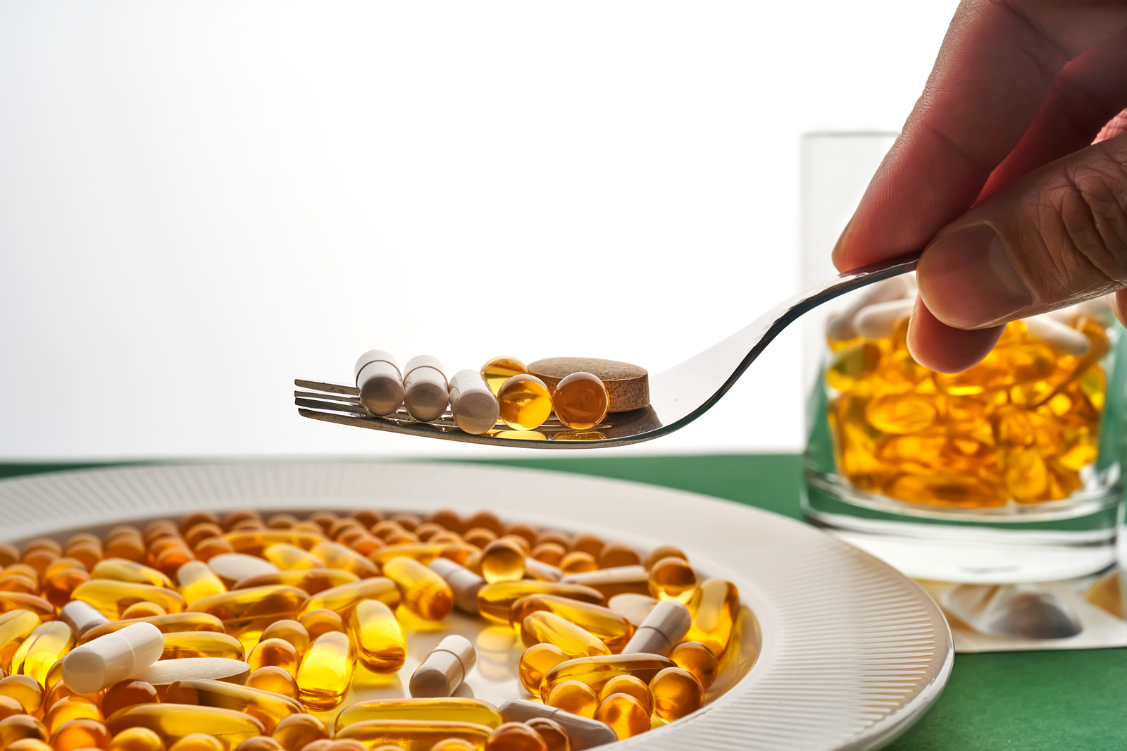 A fork holding various dietary supplements, including soft gel capsules, tablets, and pills, with a plate and glass filled with vitamins in the background, representing quality production by Softgel Manufacturing Companies