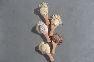 Assorted protein powders in wooden spoons displayed on a textured gray background, representing different formulations used in protein powder contract manufacturing