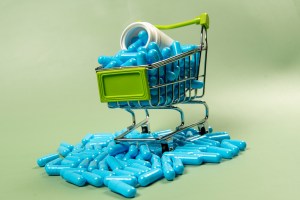 Mini shopping cart filled with blue capsules and a spilled white supplement bottle on a green background – concept for “shop all nature,” “Nature Made vitamins,” and websites to buy supplements online.