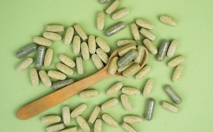A collection of natural vitamin supplements, including green tablets and capsules, scattered on a green background with a wooden spoon, symbolizing the process of vitamin manufacturing using plant-based ingredients.