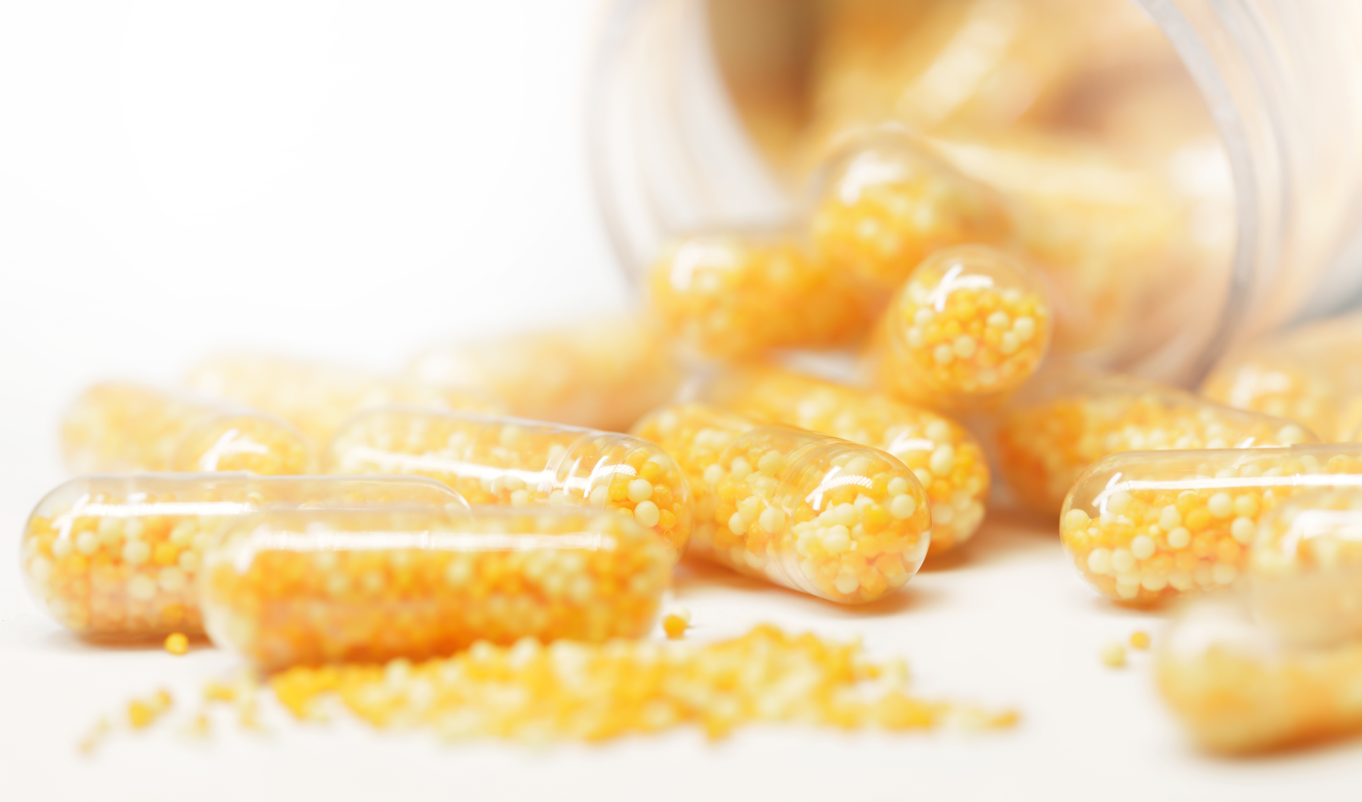 Close-up of transparent gelatin capsules filled with yellow and white vitamin granules, spilling from a bottle onto a white surface