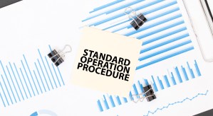 Sticky note with “Standard Operation Procedure” on top of printed graphs and charts – representing compliance in manufacturing, the importance of compliance in the manufacturing sector, and overcoming manufacturing compliance challenges.