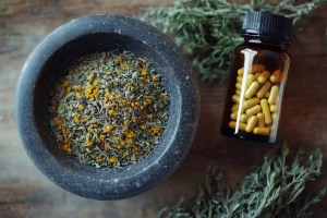 Top view of a black stone bowl filled with dried herbal leaves and flowers next to a brown supplement bottle with yellow capsules, representing herbal supplement manufacturing, organic capsule production, and natural ingredient suppliers.