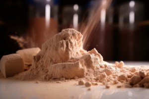 Fine beige powder being poured into a pile on a smooth surface during capsule supplement manufacturing, with compressed blocks of powder visible in the background.